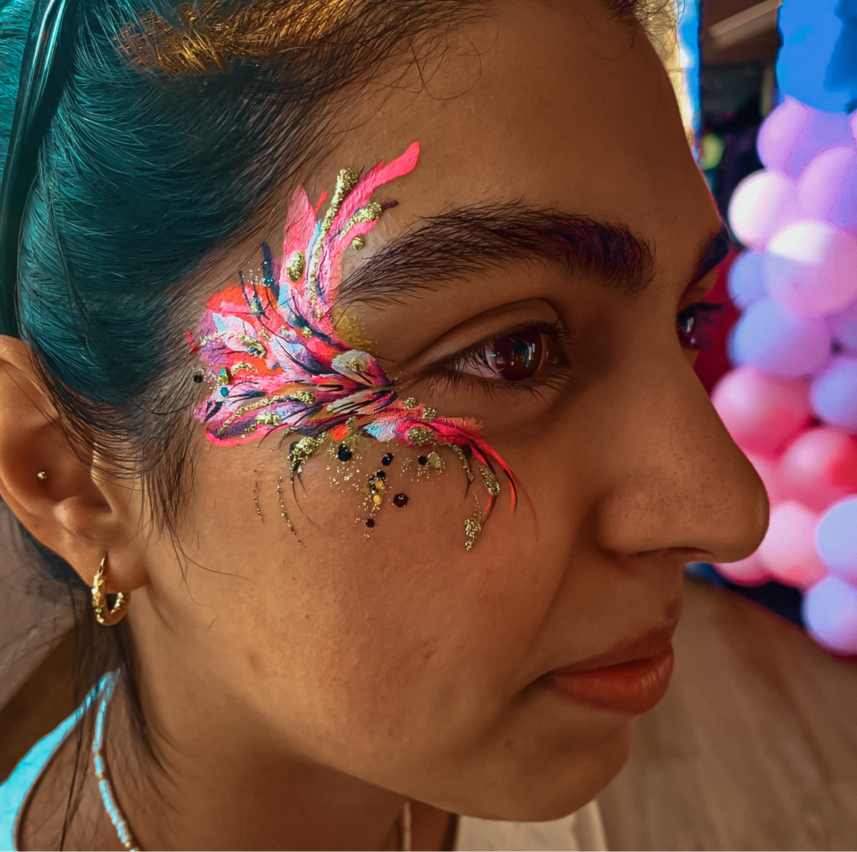 A close-up side profile of a woman with vibrant, colorful temporary face paint on her temple featuring pink, blue, gold, and black glitter details, set against a background of blurred pink and blue balloons.