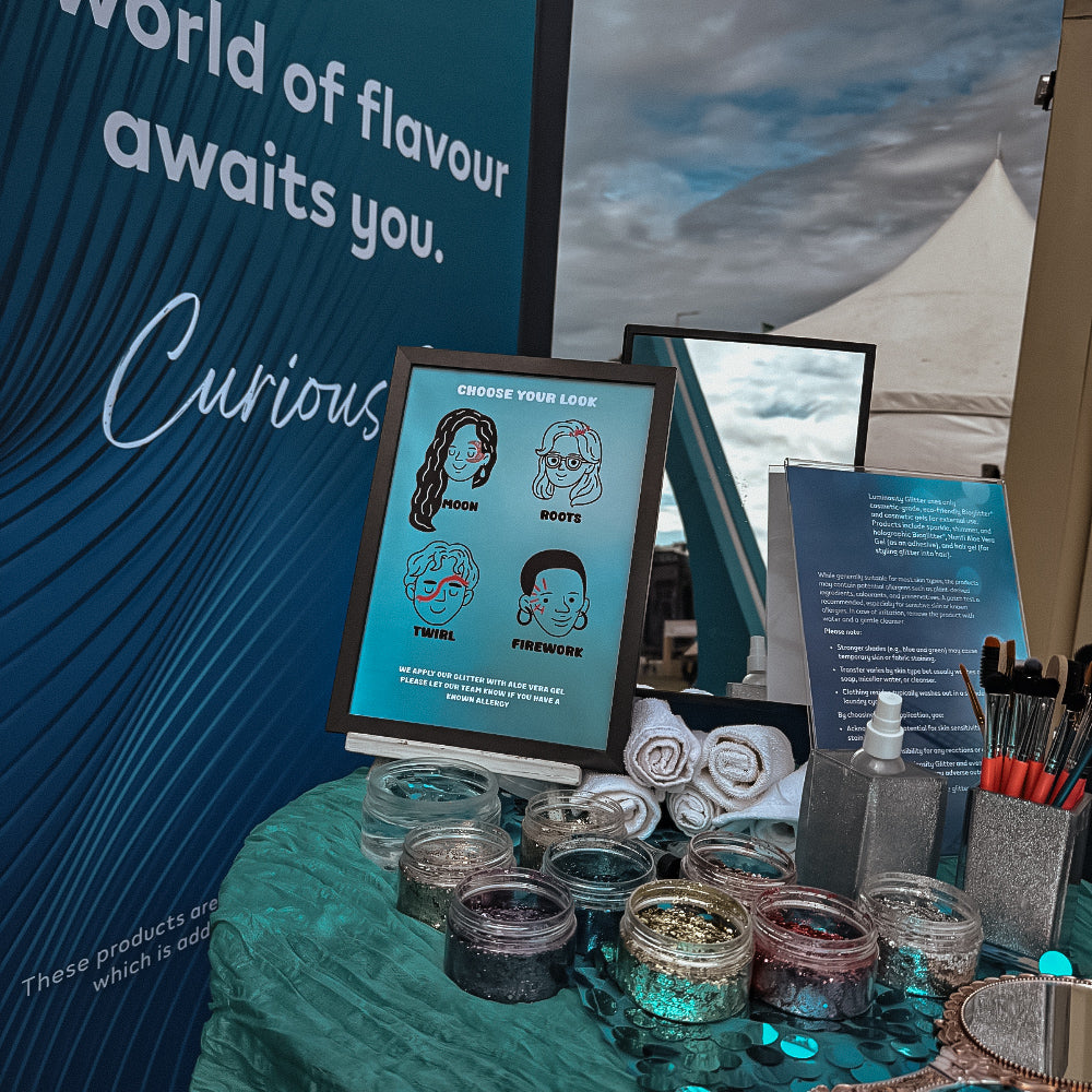 A booth display at an event featuring a digital screen showing face paint options, surrounded by jars of glitter, makeup brushes, rolled white towels, and a blue backdrop with text.