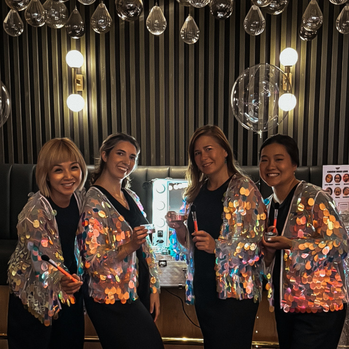Four women standing together in a dimly lit indoor setting, wearing shiny iridescent sequin jackets over black tops. The background features vertical wood paneling, hanging glass ornaments, and warm wall lights.