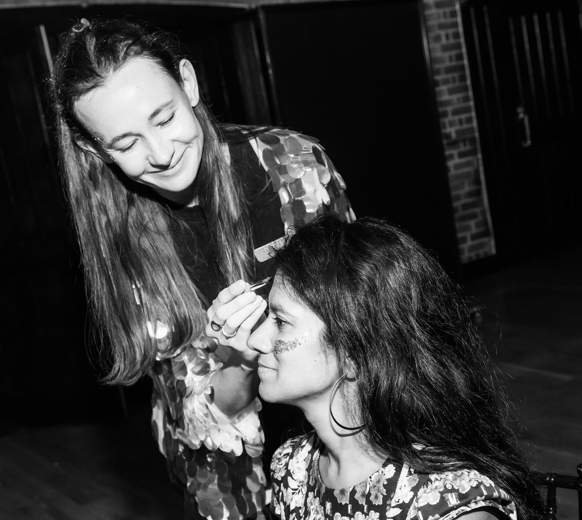 Black and white photo of a woman applying makeup to another woman's face in a dim indoor setting.