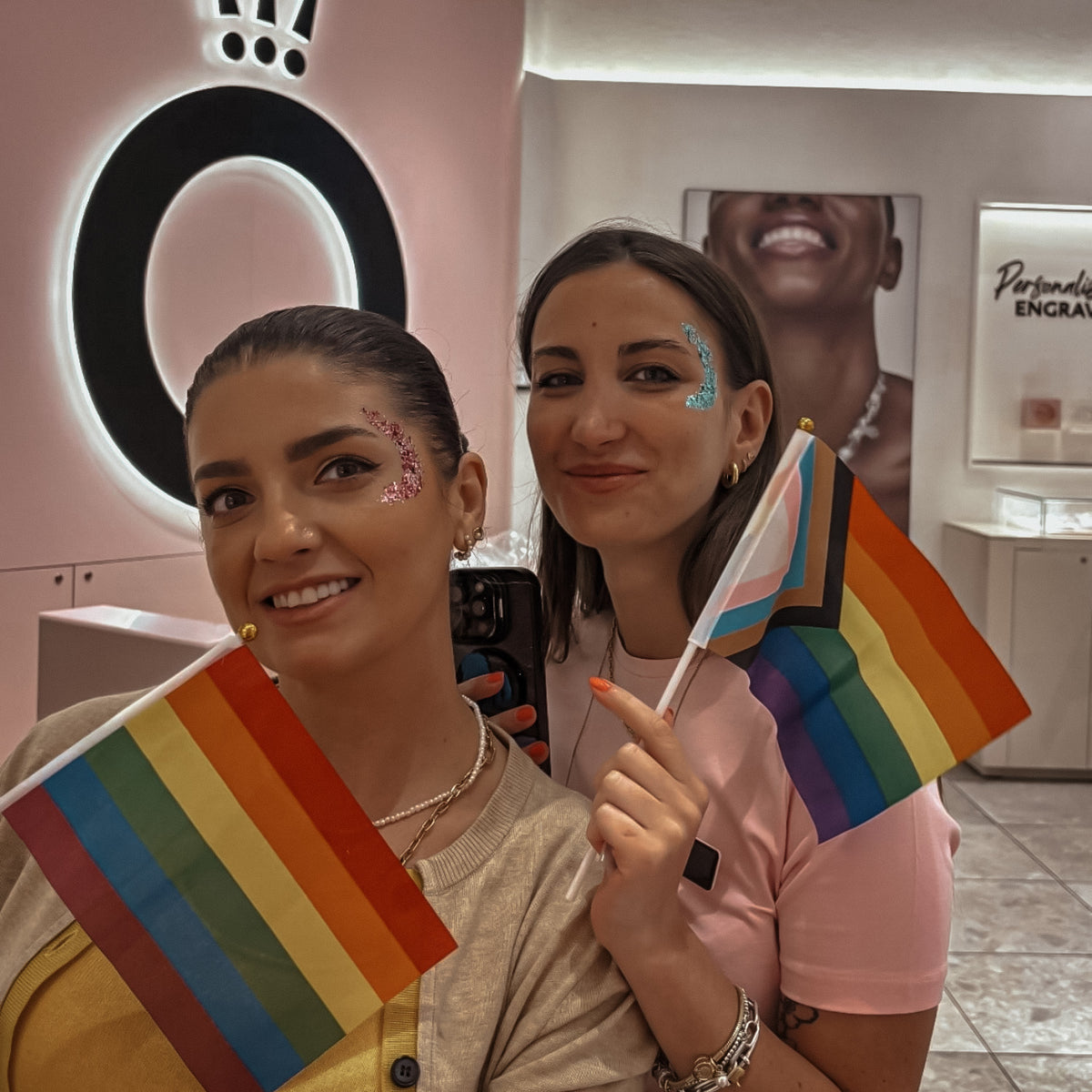 Two women smiling in a boutique setting, each holding a small rainbow pride flag. The woman in front wears a beige top and has pink glitter on her face, while the woman behind her wears a pink top and has blue glitter on her face.